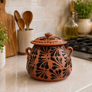 May include: A round terracotta pot with a matching lid, featuring two small side handles and a hand-painted black floral pattern across the exterior. The vessel has a wide, bulbous body and a smooth, matte finish, showcasing a traditional clay color.