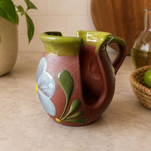 Könnte beinhalten: Ein handgefertigter, brauner Keramikkrug mit grünem Rand und Griff. Der Krug hat ein gemaltes blau-weißes Blumendesign mit grünen Blättern. Der Krug wird wahrscheinlich zum Servieren von Getränken verwendet.