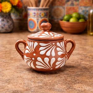May include: A terracotta sugar bowl with a lid and two handles. The bowl is decorated with white floral patterns. The background includes a vase of flowers, a bowl of limes, and other kitchen items.