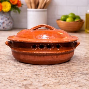 May include: A round, terracotta-colored cooking pot with a lid. The pot has a handle on the lid and two side handles. It features decorative holes with white dots. The pot is on a countertop, suggesting kitchen use.