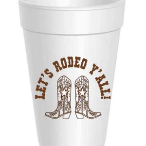 May include: A white foam cup with a brown graphic that says "Let's Rodeo Y'all!" The graphic features two cowboy boots with star details. The cup is designed for beverages.