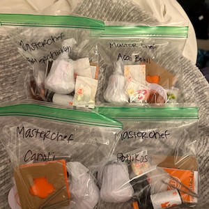 May include: Four clear plastic zip bags filled with food items. Each bag is labelled "Masterchef" with a different food item name: Superchicken, Acai Bowl, Canoli, and Potjikos. The bags are on a bed.