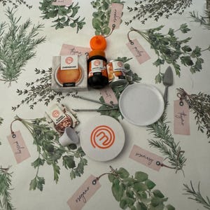 May include: A white table setting with a white plate, a white plate with a MasterChef logo, a small white bowl, a small packet of sliced almonds, a small jar of apricot jam, a small tin of apricot jam, a small bottle of apricot jam, and a small packet of cheesecake mix. The table setting is on a white background with drawings of herbs and their names.