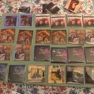 May include: A collection of square music album covers arranged in rows on a flat surface. The covers feature various artists including Steppenwolf, The Troggs, and The Mamas & The Papas, with colorful graphic designs, band photography, and printed text titles.