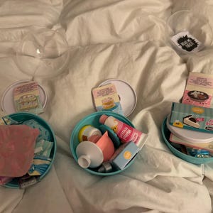 Könnte beinhalten: Drei transparente Plastikbehälter mit Deckeln, die jeweils verschiedene Bastelartikel enthalten. Die Behälter befinden sich auf einer weißen Oberfläche. Ein Behälter enthält rosa und blaue Bastelartikel, ein anderer einen weißen und blauen Kuchen und der dritte eine weiße und braune Torte.