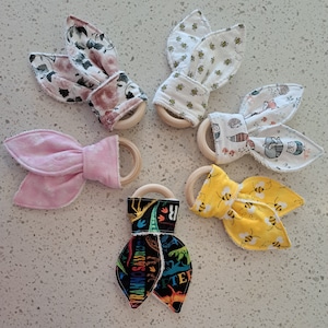 May include: Five wooden teethers with fabric coverings. The teethers are in the shape of a ring. The fabric coverings are different patterns, including floral, pink polka dots, yellow with bees, black and white dinosaurs, and a white and gray pattern with a woodland animal theme.