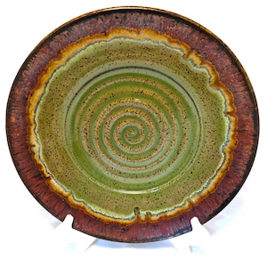 May include: A round ceramic bowl featuring a central spiral pattern in shades of olive green and brown. The interior surface is speckled with dark brown dots, while the wide, flat rim is finished in a deep, textured reddish-brown glaze.