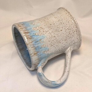 May include: A handmade ceramic mug with a speckled white glaze. The rim and handle are white, while the lower portion features a light blue and brown glaze. The mug has a large, curved handle.