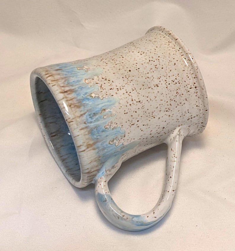 May include: A handmade ceramic mug with a speckled white glaze. The rim and handle are white, while the lower portion features a light blue and brown glaze. The mug has a large, curved handle.