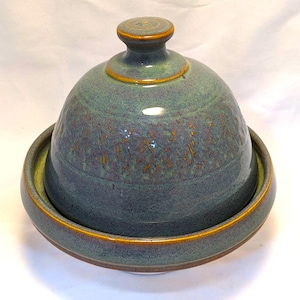 May include: A ceramic butter dish with a dome-shaped lid and a matching base. The piece features a mottled blue-green glaze with textured, brownish-orange accents across the surface. The lid has a small, rounded knob handle at the top.