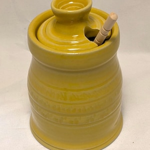May include: A yellow ceramic honey pot with a lid and a small wooden honey dipper. The pot has horizontal ridges and a rounded shape. The lid has a small opening for the dipper.