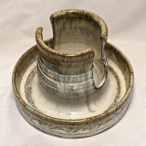 May include: A handmade ceramic spoon rest and holder. The piece features a circular base with a raised, curved holder. The colors include shades of white, brown, and blue, with a textured finish. The rim of the base has a decorative pattern.