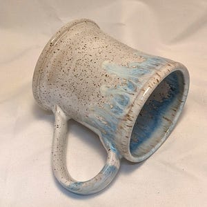 May include: Handmade ceramic mug with a speckled, off-white exterior and a blue and brown interior and rim. The mug has a curved handle and a wide opening, perfect for coffee or tea. The mug is approximately 10 cm tall.