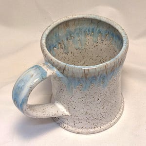 May include: A handmade ceramic mug with a speckled white exterior and a light blue interior and handle. The rim features a dripped blue glaze. The mug is designed for hot beverages.