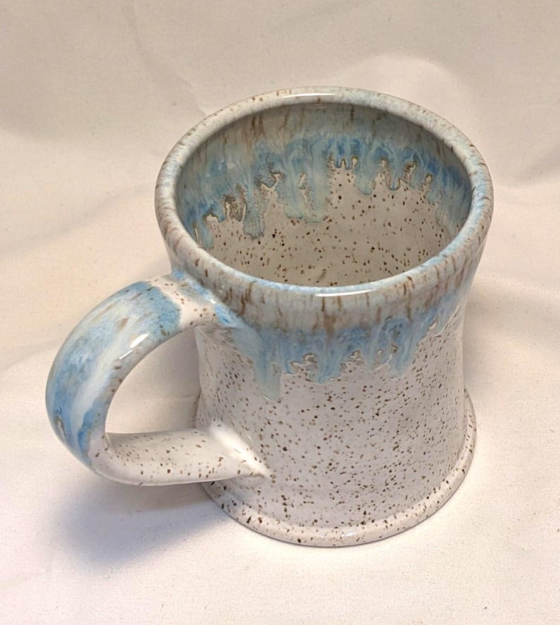 May include: A handmade ceramic mug with a speckled white exterior and a light blue interior and handle. The rim features a dripped blue glaze. The mug is designed for hot beverages.