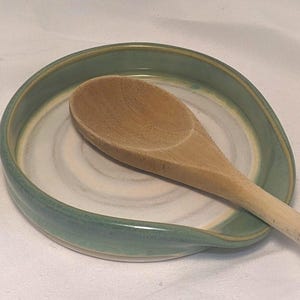 May include: A handmade ceramic spoon rest with a light green rim and a white and beige interior. A wooden spoon rests on the spoon rest. The spoon rest is designed to hold a spoon while cooking.