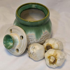 May include: A handmade ceramic garlic keeper with a green and cream glaze. The lid has a small knob and several holes. Three heads of garlic are next to the keeper. The keeper is a kitchen storage item.