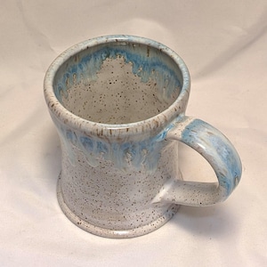 May include: A handmade ceramic mug with a speckled white base and a light blue glaze around the rim and handle. The mug has a sturdy handle and a slightly flared shape. The interior is a speckled white colour.