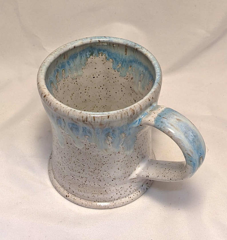 May include: A handmade ceramic mug with a speckled white base and a light blue glaze around the rim and handle. The mug has a sturdy handle and a slightly flared shape. The interior is a speckled white colour.