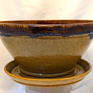 May include: A ceramic planter bowl with a speckled tan finish and a wide, dark brown glazed rim. The bowl sits securely on a matching circular saucer, which features the same speckled tan texture and a raised edge to contain excess water drainage.