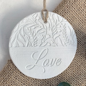 May include: A white, circular ornament featuring a botanical relief design and the word "Love". It has a jute twine loop for hanging. The background is a hessian-style fabric.