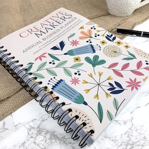 May include: A floral-patterned business planner with the title "CREATIVE MAKERS ANNUAL BUSINESS PLANNER." The spiral-bound notebook features a design with blue, pink, and green flowers. A pen and a cup are visible in the background.