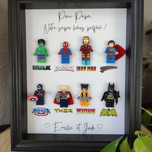 Peut inclure: Une œuvre d'art encadrée avec des figurines Lego de super-héros, dont Hulk, Spider-Man, Iron Man et Superman. L'œuvre comprend le texte "Pour Papa, Notre super héros préféré !" et les noms "Emilie et Jade".