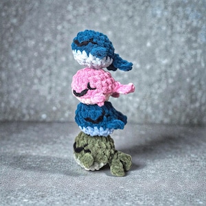 Könnte beinhalten: Ein Stapel von vier gehäkelten Wal-Plüschtieren in Blau-, Rosa- und Grüntönen. Jeder Wal hat ein lächelndes Gesicht mit geschlossenen Augen und einen kleinen Schwanz. Die Plüschtiere sind übereinander gestapelt.