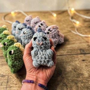Könnte beinhalten: Eine Sammlung handgefertigter gehäkelter Dinosaurier-Spielzeuge in verschiedenen Pastellfarben, darunter Grün, Blau und Lila. Die Dinosaurier haben kleine, gestickte Details und sind auf einer Holzoberfläche angeordnet. Eine Hand hält einen hellblauen Dinosaurier im Vordergrund.