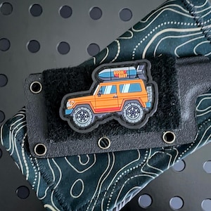 以下が含まれることがあります： 「BOOM BABY」と書かれたサーフボードを載せたオレンジ色のカートゥーン風のジープが描かれたベルクロパッチが付いた黒いナイフシースです。