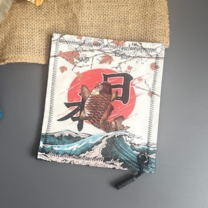 May include: A small, rectangular pouch featuring a Japanese-inspired design. The artwork depicts a koi fish leaping from ocean waves against a red sun, with Japanese characters. The pouch has a black zipper and a black pull.