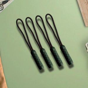 May include: A set of four dark green zipper pulls, each featuring a textured, diamond-patterned cylindrical grip attached to a thin, black braided cord loop. These functional accessories are designed to be attached to zippers for easier handling.