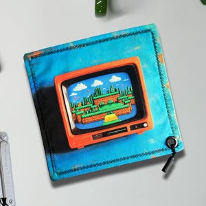Puede incluir: Una alfombrilla de ratón cuadrada azul con un gráfico de televisión naranja retro que muestra una escena de videojuego pixelada. La alfombrilla tiene un borde cosido y un cordón negro sujeto a una arandela.