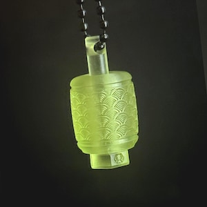 May include: A glowing, light green pendant necklace with a cylindrical, lantern-like charm. The charm features a detailed, wave-like pattern and is suspended from a black beaded chain. The background is a dark, solid color.