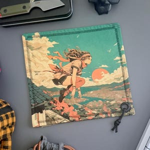 以下が含まれることがあります： 夕焼けの街並みを飛び越えるアニメ風イラストが描かれた正方形の布製品。画像には、ターコイズブルーの空、オレンジ色の太陽、黒いステッチの入った詳細なボーダーがあります。黒いリングとコードが取り付けられています。