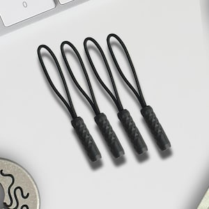 Puede incluir: Cuatro tiradores de cremallera negros con agarres texturizados y cordones en bucle. Los tiradores están dispuestos en fila, con un teclado y otros objetos en el fondo. Probablemente son para ropa o bolsos.