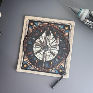 May include: Square beige coaster featuring a detailed illustration of the Millennium Falcon spaceship. The artwork is in shades of blue, brown, and white, with a black stitched border. A small black loop is attached to the coaster.