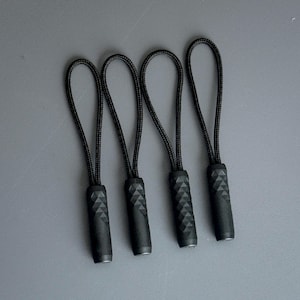 May include: Four black zipper pulls with textured, geometric designs on the pull tabs. Each pull has a loop made of black cord. The pulls are arranged in a row against a gray background.