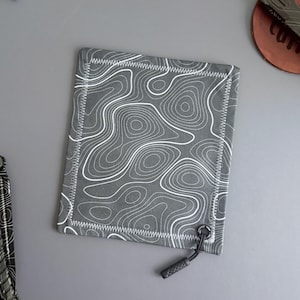 May include: A gray fabric pouch with a white topographic map pattern. The pouch has a white zigzag stitch border and a black cord with a plastic end. A knife and a coaster are partially visible in the background.