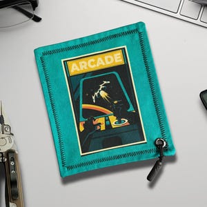 May include: Teal fabric pouch with a retro arcade game design. The pouch features a black zipper pull and a stitched border. The arcade graphic shows a spaceship and the word "ARCADE" in yellow.