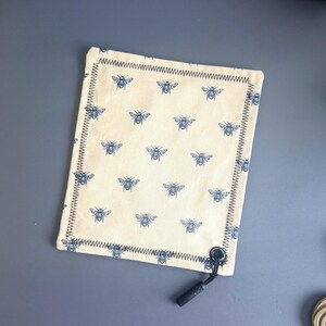 May include: Cream-colored rectangular fabric item with a repeating pattern of blue bees. The edges are finished with a dark blue zigzag stitch. A black cord with a loop and a small black tassel is attached to one corner.