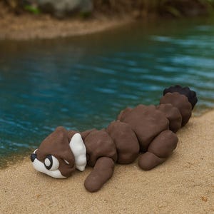 May include: A brown, white, and black sculpture of a stylized animal, possibly a river otter, lies on a sandy beach near water. The sculpture has a textured surface and is positioned near the water's edge, with green reeds in the background.