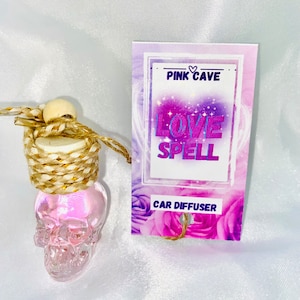 Puede incluir: Difusor de coche Pink Cave Love Spell en una botella de cristal transparente con forma de calavera con un cordón dorado y marrón que cuelga de una cuenta de madera. La botella está sobre una superficie blanca. La etiqueta dice "Pink Cave Love Spell Car Diffuser".