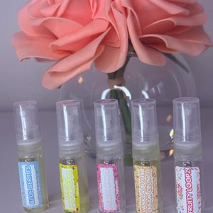 以下が含まれることがあります： 「Blue Berries」、「Lemon Poundcake」、「Peach Rings」、「Vanilla Butter Cream Crunch」、および「Fruity Loopz」と書かれたラベルが付いた5つの小さな香水スプレーボトル。大きなピンク色のバラが、背景の透明なガラスの花瓶に入っています。