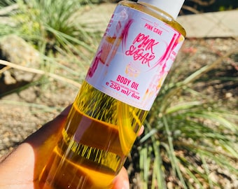 Może przedstawiać: Przezroczysta butelka olejku do ciała Pink Sugar z białym dozownikiem z pompką. Etykieta zawiera różowy i biały tekst oraz grafikę. Butelka zawiera 250 ml olejku.