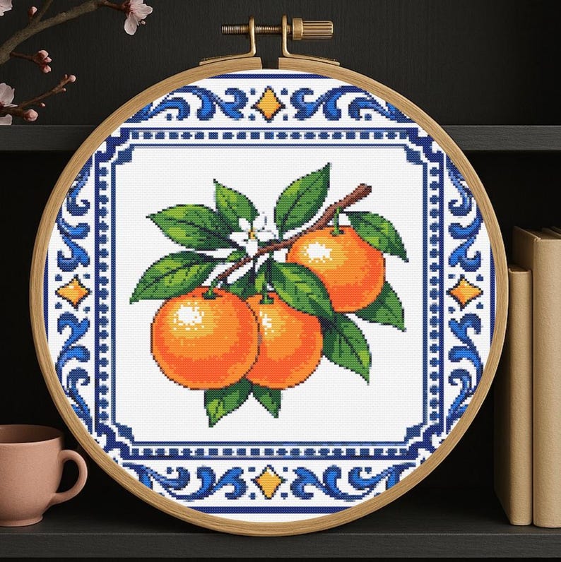 May include: Cross-stitch embroidery of oranges on a branch with green leaves and white flowers, set in a wooden hoop. The design is framed by a blue and white decorative border.