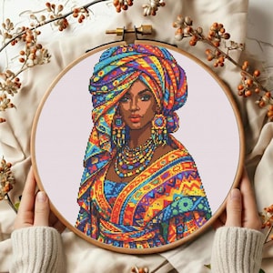 May include: A cross-stitch portrait of a woman wearing a colorful head wrap, earrings, and necklace. The artwork is framed in a wooden embroidery hoop. The woman's skin is brown, and the wrap and clothing feature a vibrant mix of blue, orange, yellow, and green.