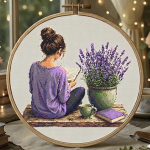 May include: A circular wooden embroidery hoop containing a cross-stitch pattern of a person with a brown hair bun wearing a purple long-sleeve shirt, sitting next to a potted lavender plant, a small green cup, and a closed purple book on a wooden surface.