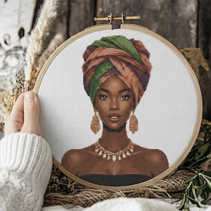 May include: Cross-stitch portrait of a woman wearing a colorful head wrap, gold earrings, and a necklace. The embroidery is framed in a wooden hoop. The woman has brown skin and is wearing a black top.