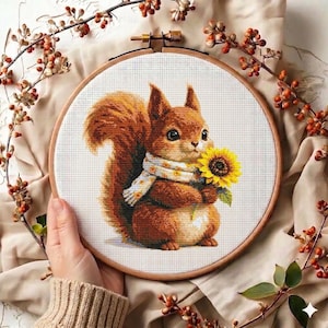 May include: Cross-stitch embroidery of a squirrel holding a sunflower, wearing a scarf with a floral pattern. The squirrel is brown with a fluffy tail, set in a wooden embroidery hoop. The background is a neutral fabric with decorative branches.